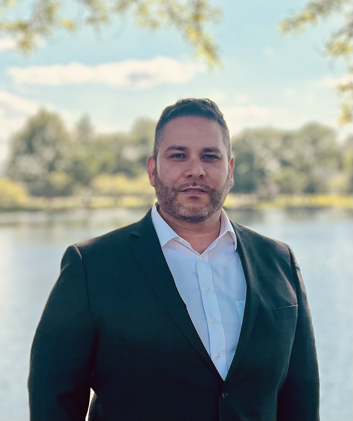 gil gonzales headshot Gil Gonzales - photo from the chest up in a gray suit jacket with the lake in the background.
