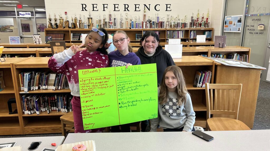 4 Barberton students standing in the library with a handmade sign listing "unhealthy" and "healthy" behavior.